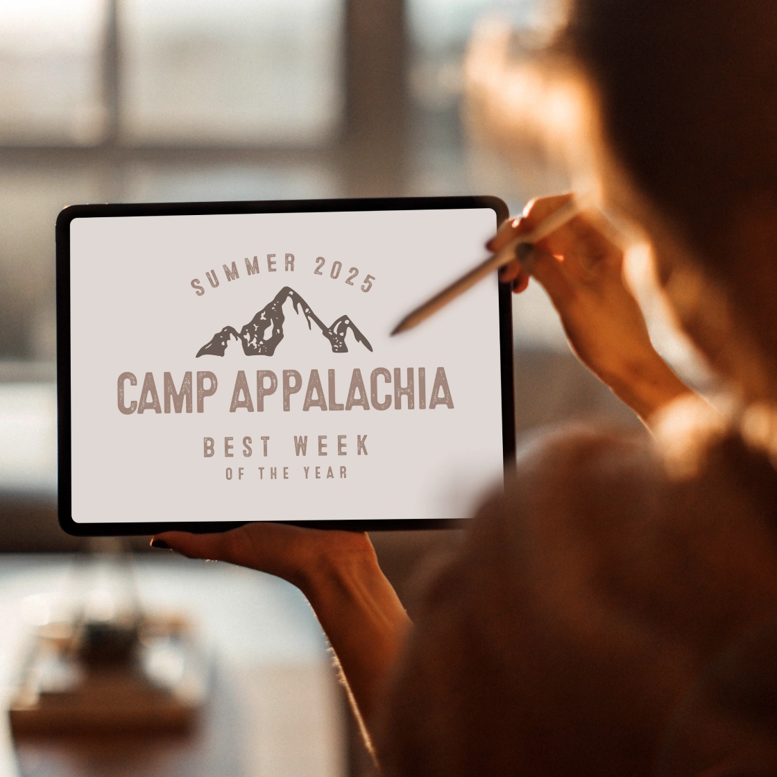 Designer holding a tablet displaying a vintage-style summer camp program logo with a mountain illustration